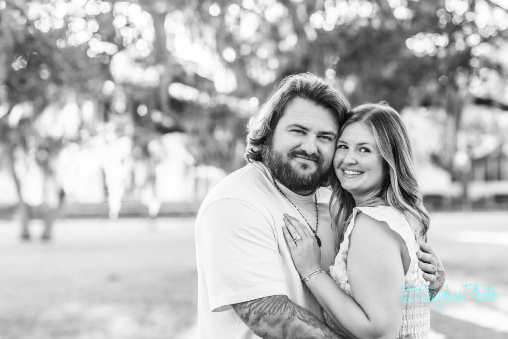 A bearded man and a woman smile while embracing outdoors, standing close together under trees. The image is in black and white, with a blurred natural background. Vero Beach Photoshoot
