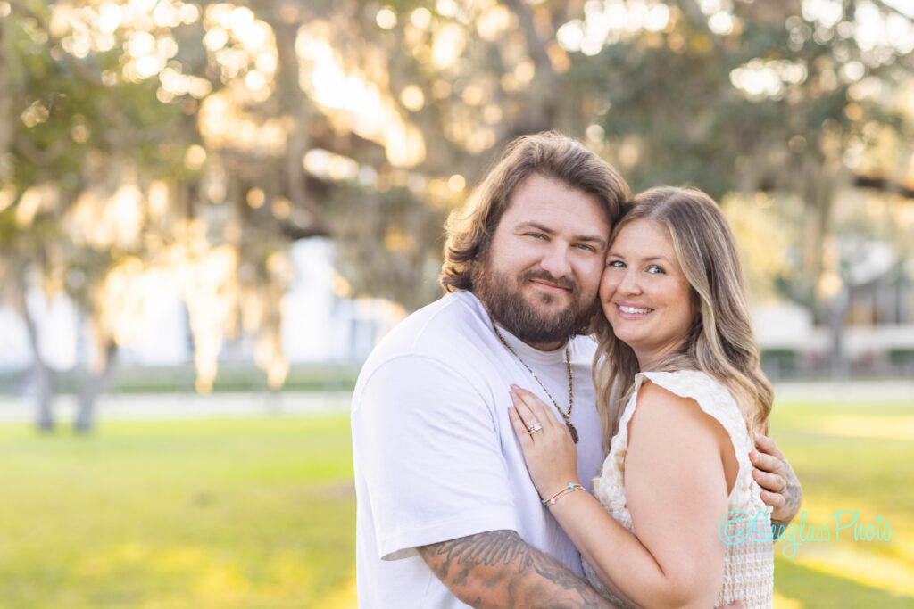 A couple stands outdoors, smiling and embracing. The man has a beard and long hair, wearing a white shirt, and the woman has long light hair and wears a light dress. Sunlight and trees are blurred in the background. Vero Beach Photoshoot