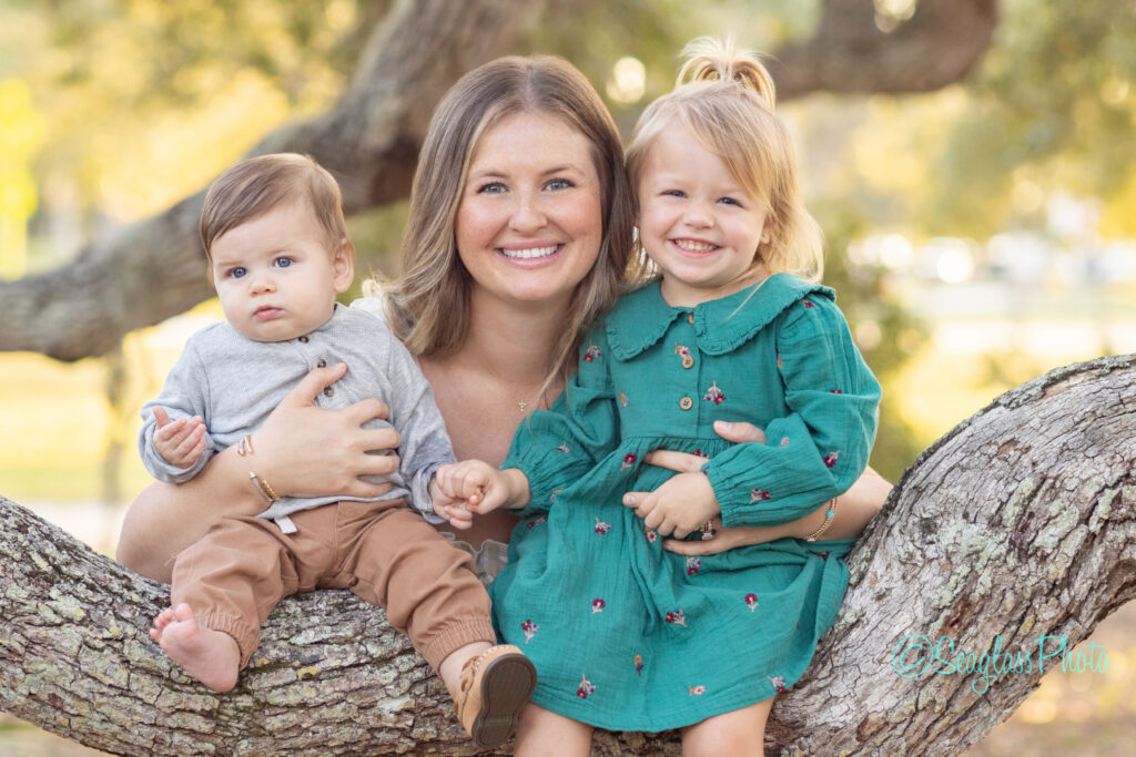 A smiling woman sits on a tree branch, holding a baby in grey and brown clothes on her left and a toddler in a green dress on her right. The background is outdoors with soft, green foliage. Vero Beach Photoshoot