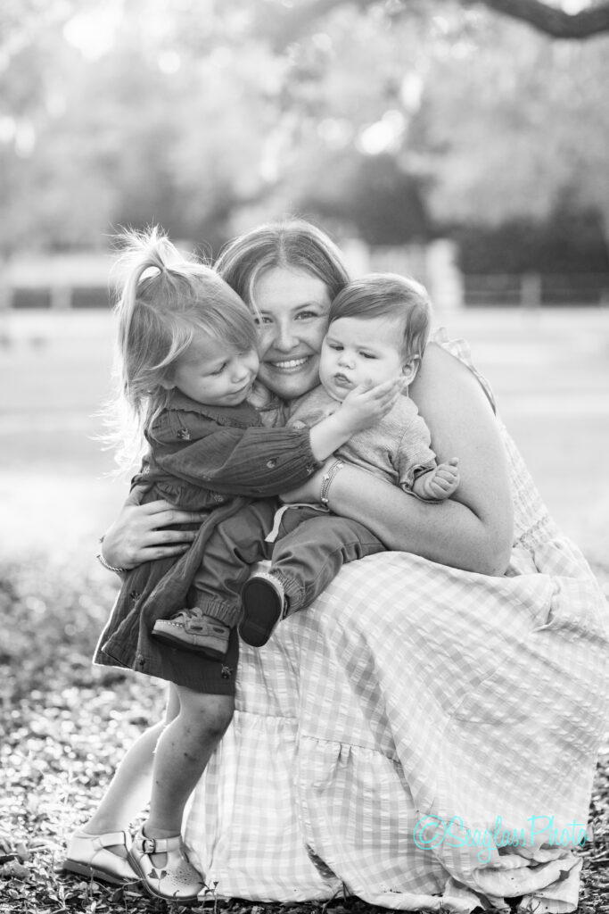 A woman in a checkered dress hugs two small children outdoors, smiling warmly. The little girl stands to her left, while the baby boy sits on her lap. Sunlight filters through the trees in the background. Vero Beach Photoshoot