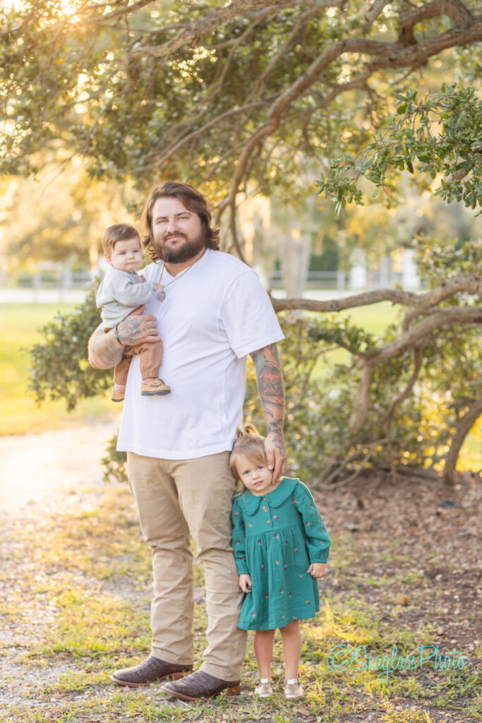A bearded man in a white shirt stands outdoors holding a young child on his arm, while another young child in a green dress stands by his side. Warm sunlight filters through trees in the background. Vero Beach Photoshoot