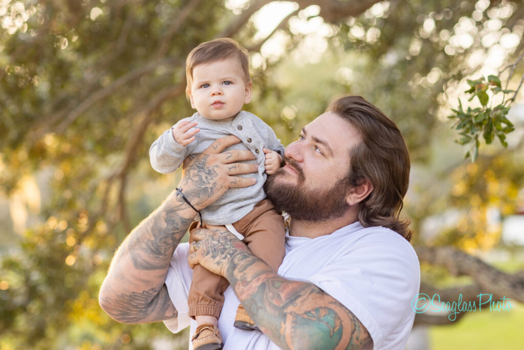 A bearded man with tattoos smiles while holding a baby in his arms outdoors. The baby, wearing a gray shirt and brown pants, looks ahead with a curious expression. Trees and sunlight are in the background. Vero Beach Photoshoot