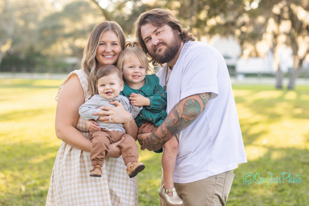 A smiling family of four poses outdoors. The mother holds a baby boy, while the father, who has tattoos, holds a young girl. Sunlight filters through trees in the background, creating a warm, cheerful atmosphere. Vero Beach Photoshoot