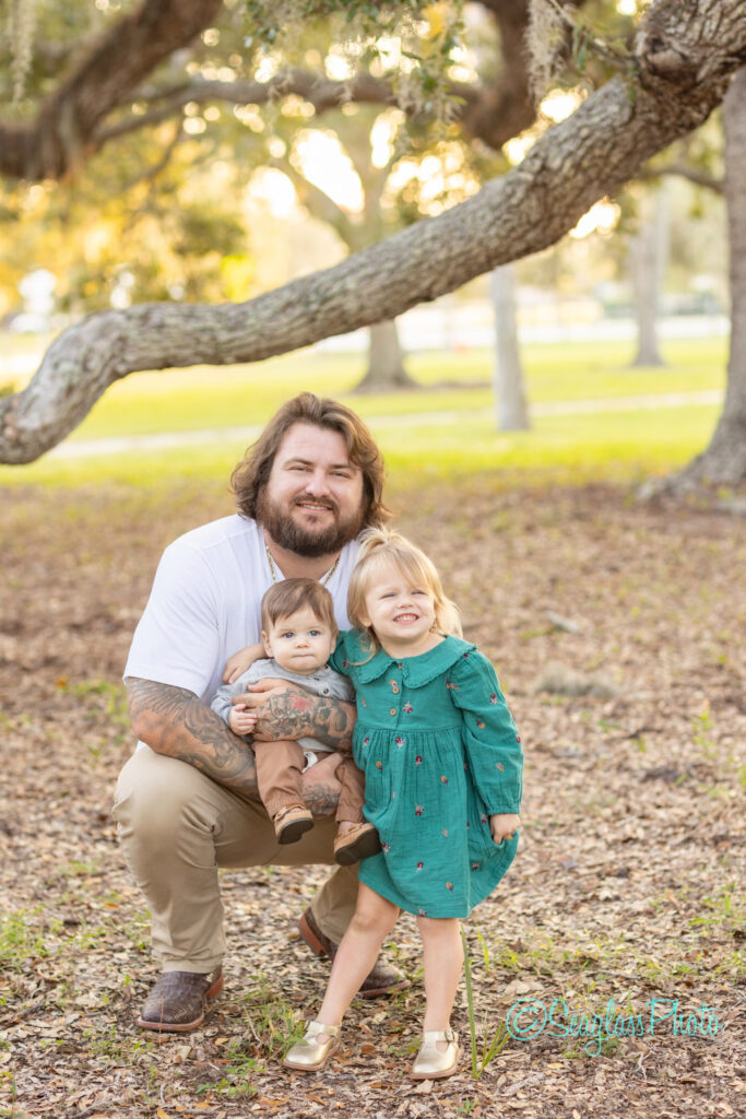 A bearded man squats outdoors under a large tree, smiling as he holds a baby. A young girl in a green dress stands beside them, also smiling. The background shows green grass and more trees. Vero Beach Photoshoot