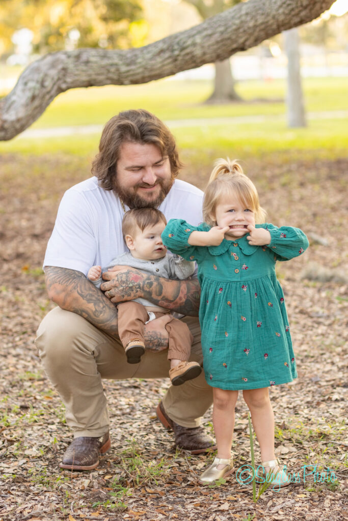 A bearded man crouches outdoors, holding a baby boy while a young girl in a green dress stands in front, playfully pulling her cheeks. They are surrounded by trees and fallen leaves. Vero Beach Photoshoot