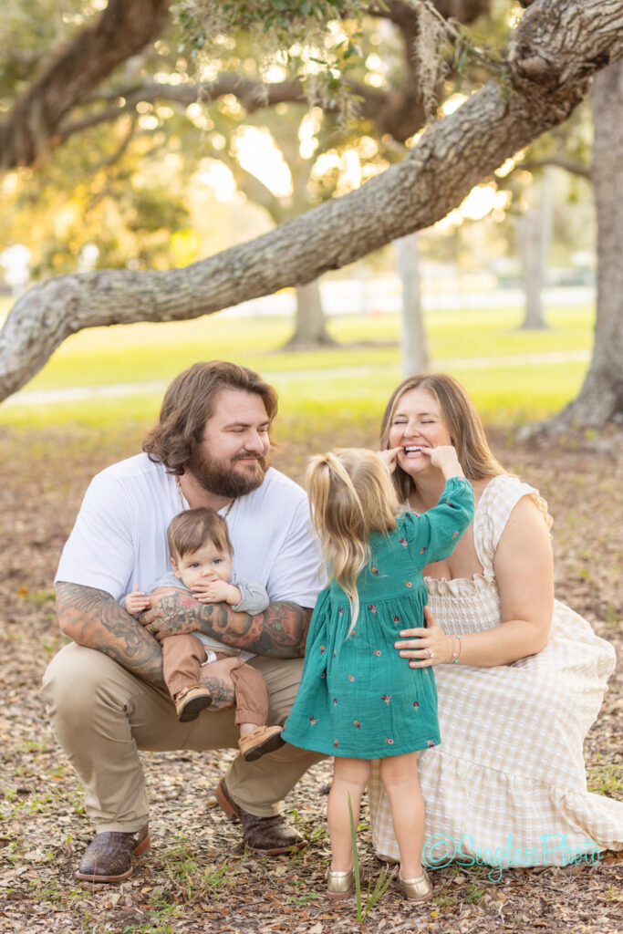 A man and woman kneel on the ground outdoors with their two young children. The girl in a green dress touches the womans face while the man smiles, holding a baby boy. Sunlight filters through the trees in the background. Vero Beach Photoshoot