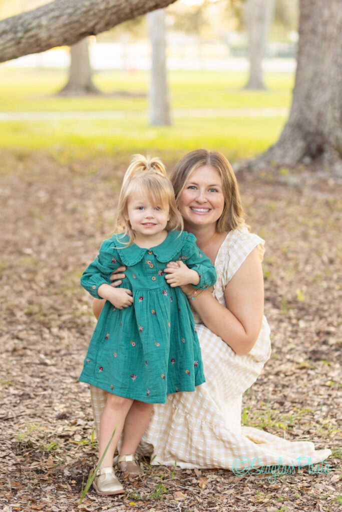 A smiling woman in a light-colored dress kneels next to a young girl in a green dress, holding her hands, outdoors in a park with trees and sunlight in the background. Vero Beach Photoshoot