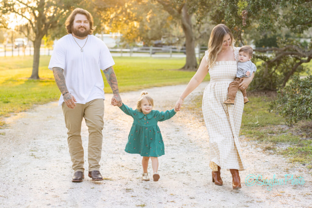 A smiling family of four walks on a sunlit path. The father, in a white shirt and jeans, holds his daughters hand; the mother, in a plaid dress, holds their toddler son. Trees and soft sunlight fill the background. Vero Beach Photoshoot