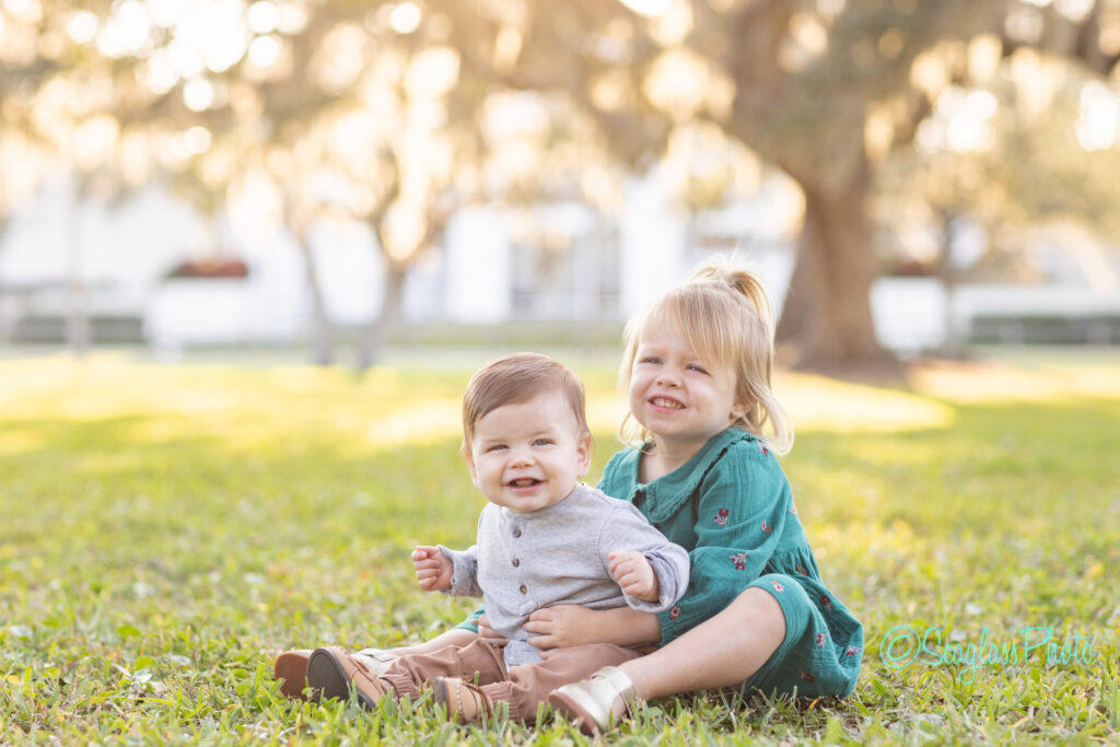 Two young children, a smiling baby boy in a gray outfit and an older girl in a green dress, sit together on grass in a sunlit park with trees in the background. Vero Beach Photoshoot