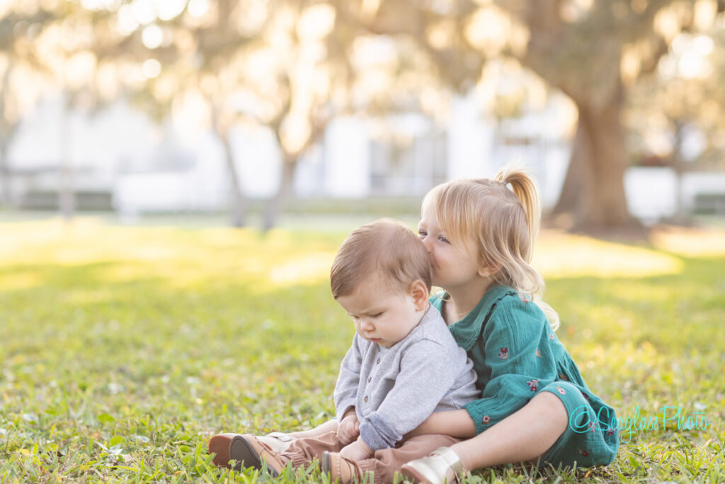 A young girl in a green dress hugs and kisses a baby boy sitting on the grass in a sunlit outdoor setting with trees and blurred background. Vero Beach Photoshoot