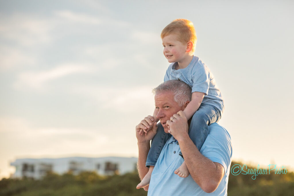 An older man carries a smiling young boy on his shoulders outdoors. Both wear light blue shirts. The background shows greenery, a white building, and a clear sky. Vero Beach Photoshoot