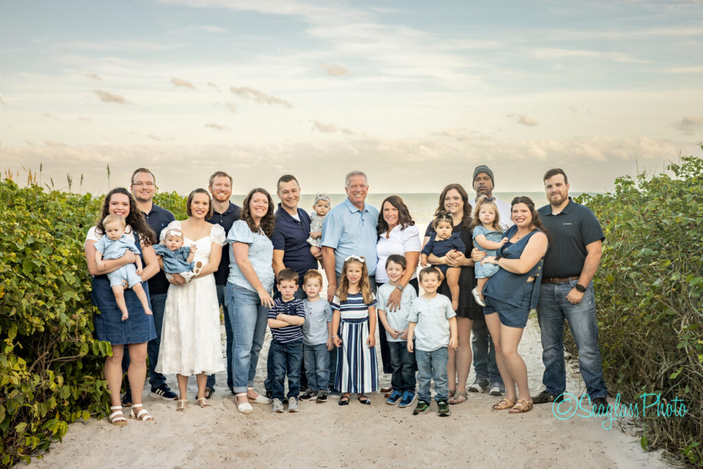 A large multigenerational family poses together on a sandy beach, surrounded by greenery and under a cloudy sky. The group includes adults, children, and babies, all dressed in blue, white, and denim clothing, smiling at the camera. Vero Beach Photoshoot