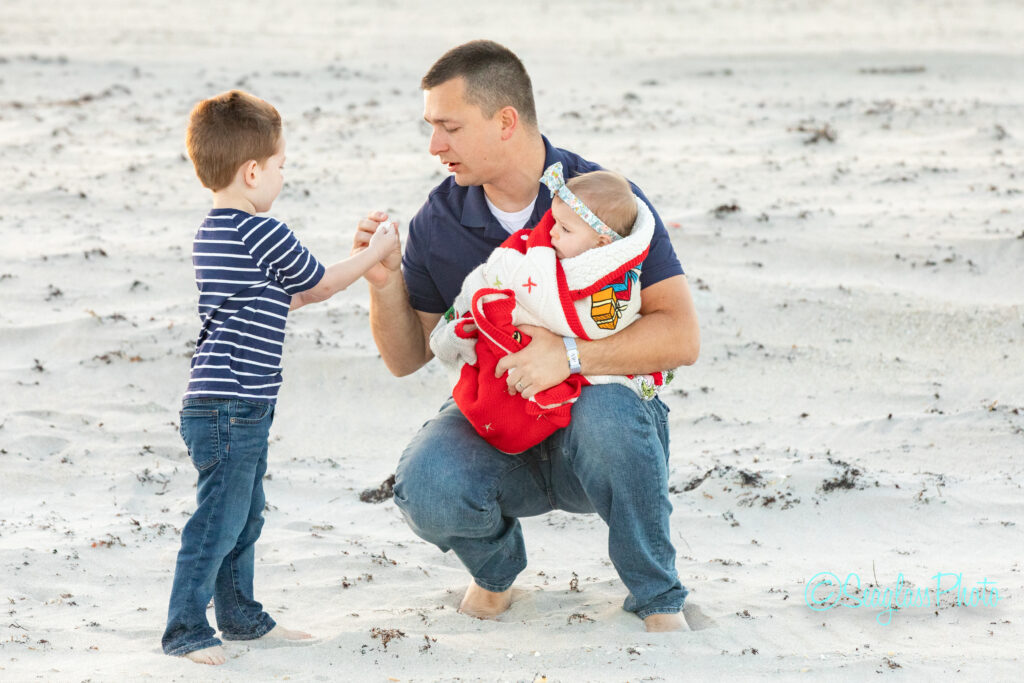A man kneels on a sandy beach, holding a baby wrapped in a red blanket while fist bumping a young boy in a striped shirt. Vero Beach Photoshoot