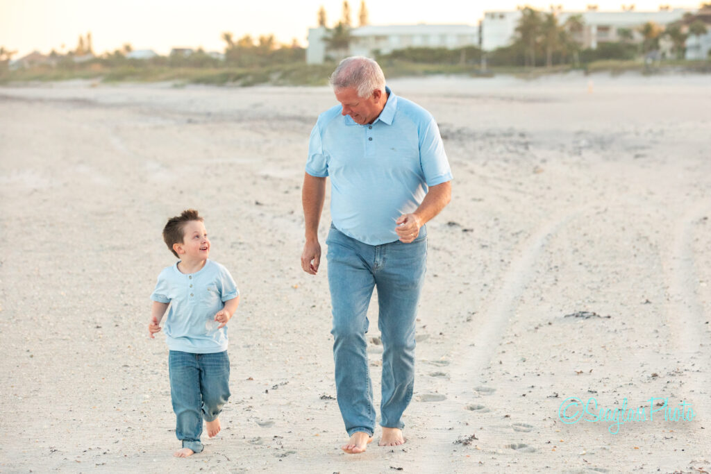 An older man and a young boy, both barefoot and wearing light blue shirts and jeans, smile as they walk together along a sandy beach with houses and trees in the background. Vero Beach Photoshoot