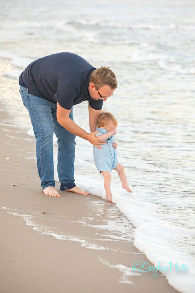 A man in a navy shirt and jeans helps a baby in a blue outfit touch the ocean waves at the shore, both barefoot on wet sand with gentle waves rolling in. Vero Beach Photoshoot