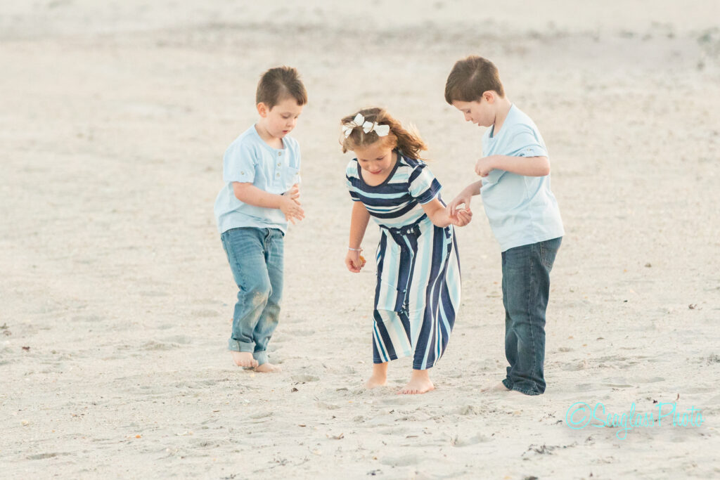 Three young children, two boys in light blue shirts and jeans and a girl in a striped dress, play together on a sandy beach, looking down at the sand. Vero Beach Photoshoot