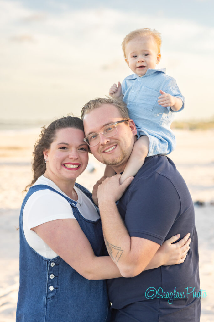 A smiling couple poses on a sandy beach, with the man holding a young child on his shoulders. The child, wearing a light blue outfit, looks at the camera while the beach and sky are softly blurred in the background. Vero Beach Photoshoot