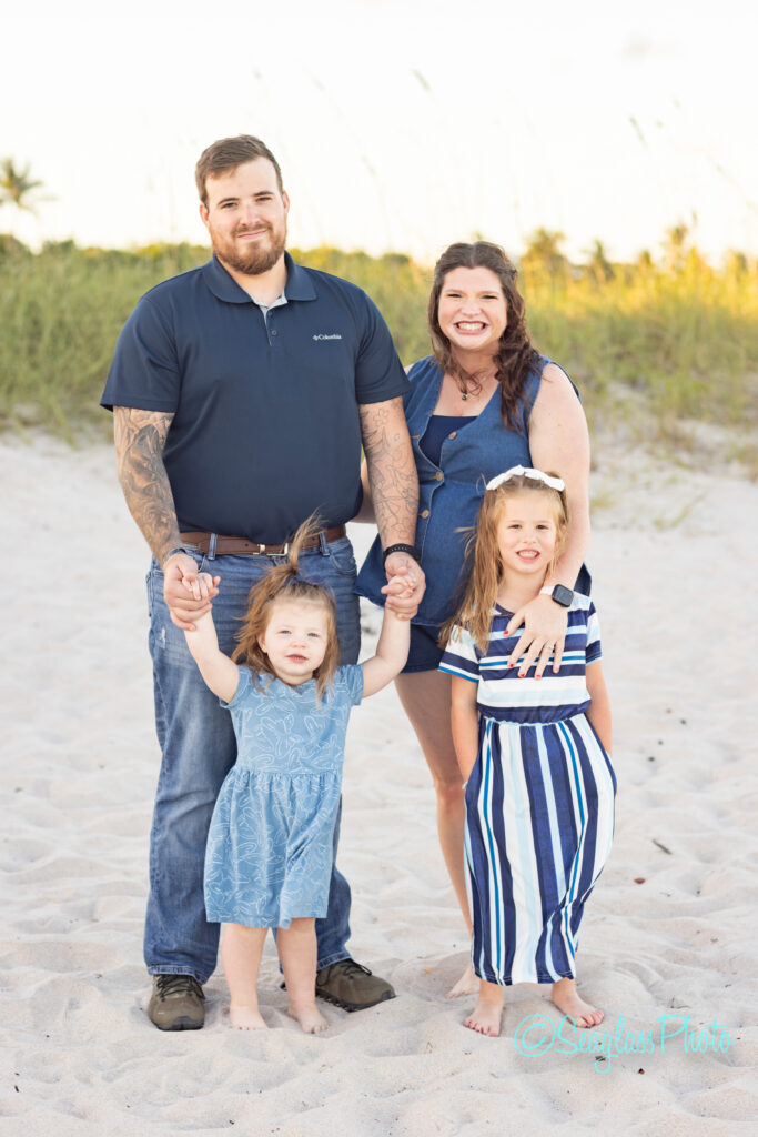 A smiling family of four poses on a sandy beach. The father, with tattoos and a beard, stands next to the mother, who is wearing a blue dress. Their two young daughters stand in front, holding hands and smiling. Vero Beach Photoshoot