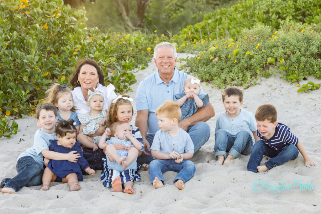 A smiling older couple sits on a sandy beach surrounded by nine young children, including several toddlers and babies. Green bushes and yellow flowers are visible in the background. Everyone appears happy and relaxed. Vero Beach Photoshoot