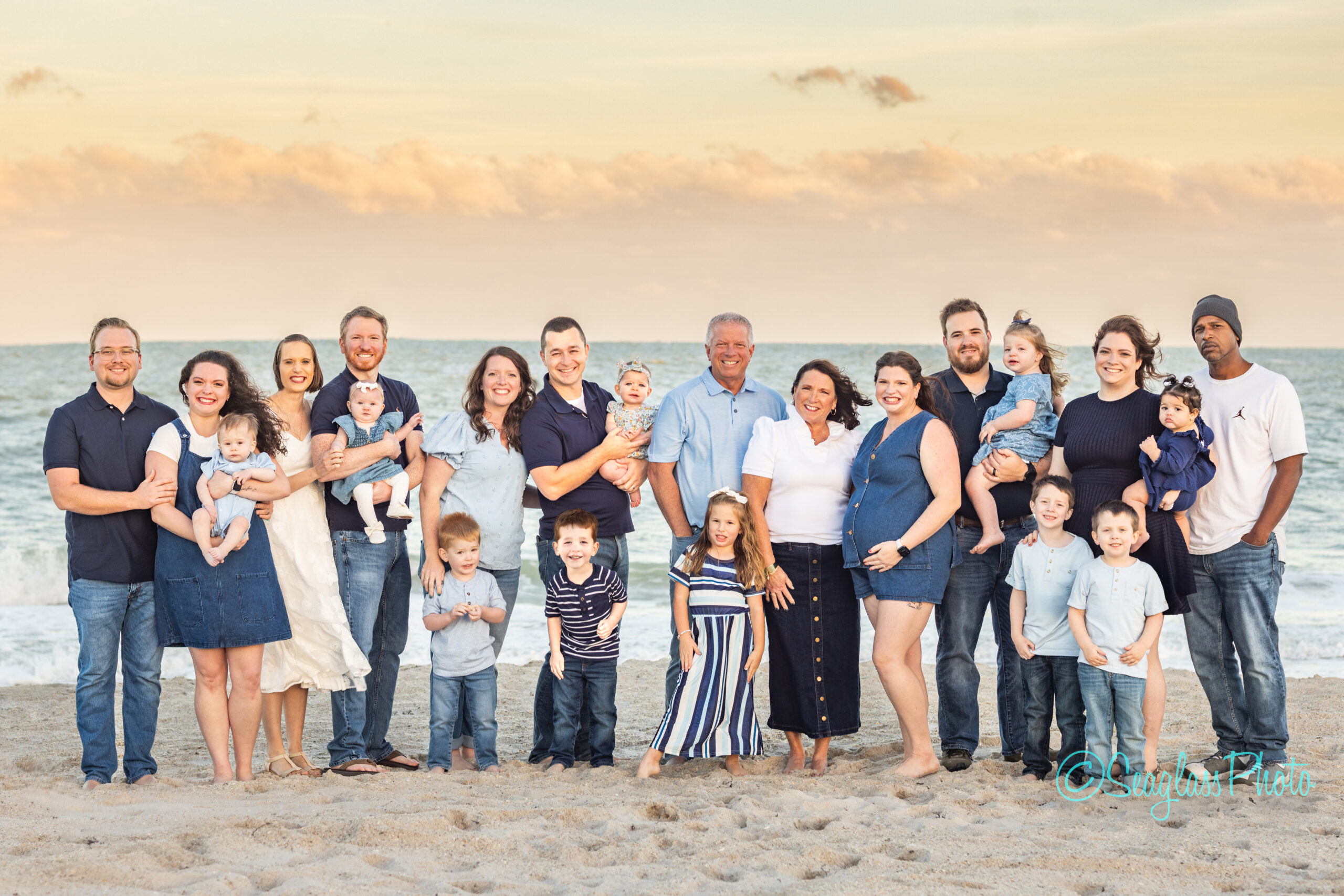 A large family of adults and children pose together on a sandy beach at sunset, smiling for the camera with the ocean and sky in the background. Vero Beach Photoshoot