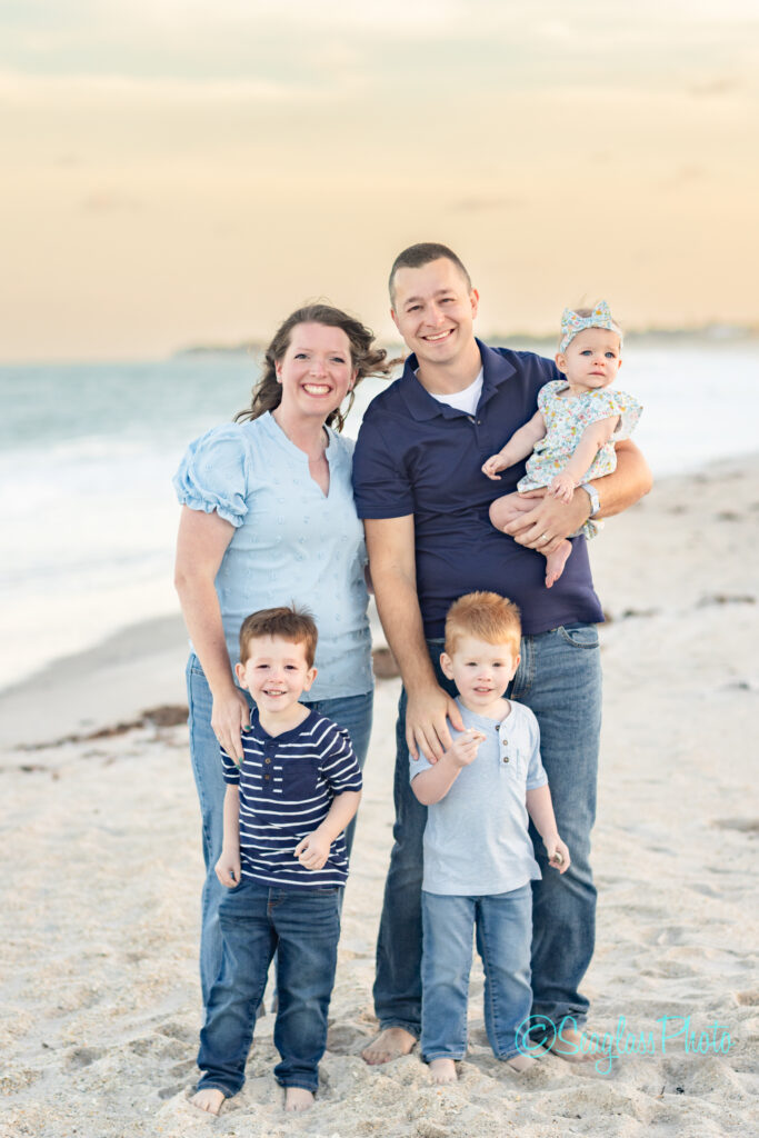 A smiling family of five stands on a sandy beach at sunset. The parents hold a baby girl, while two young boys stand in front of them. The ocean waves and sky are in the background. Vero Beach Photoshoot