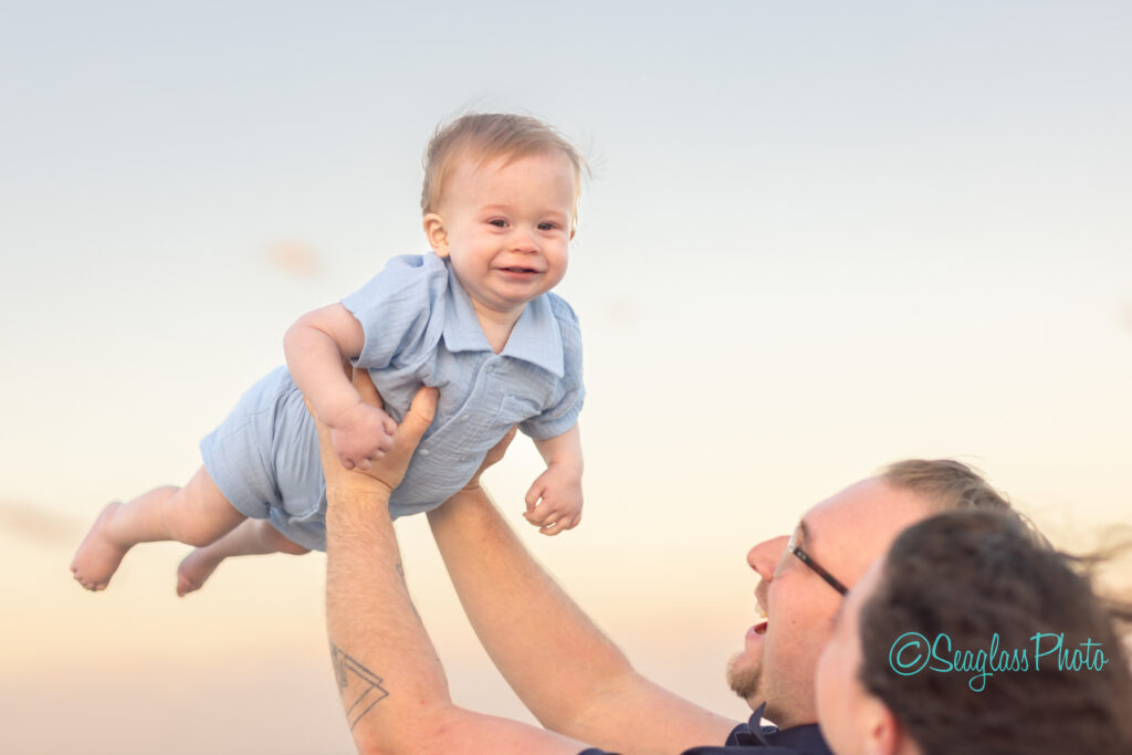 An adult lifts a smiling baby boy in a light blue outfit into the air at sunset, both appearing joyful against a soft, pastel sky. Vero Beach Photoshoot