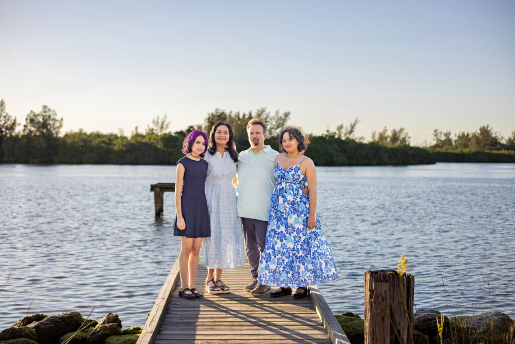 Four people stand close together on a wooden dock over a calm lake, with trees and greenery in the background under a clear sky. Three women and one man are smiling, dressed in summer clothes. Vero Beach Photoshoot