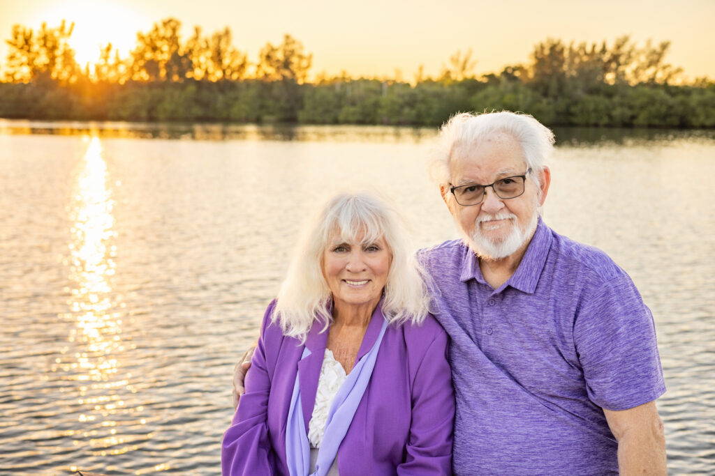 An older couple smiles while posing by a calm lake at sunset. The woman wears a purple blazer and the man wears a purple polo shirt. Trees and golden sunlight reflect on the water in the background. Vero Beach Photoshoot
