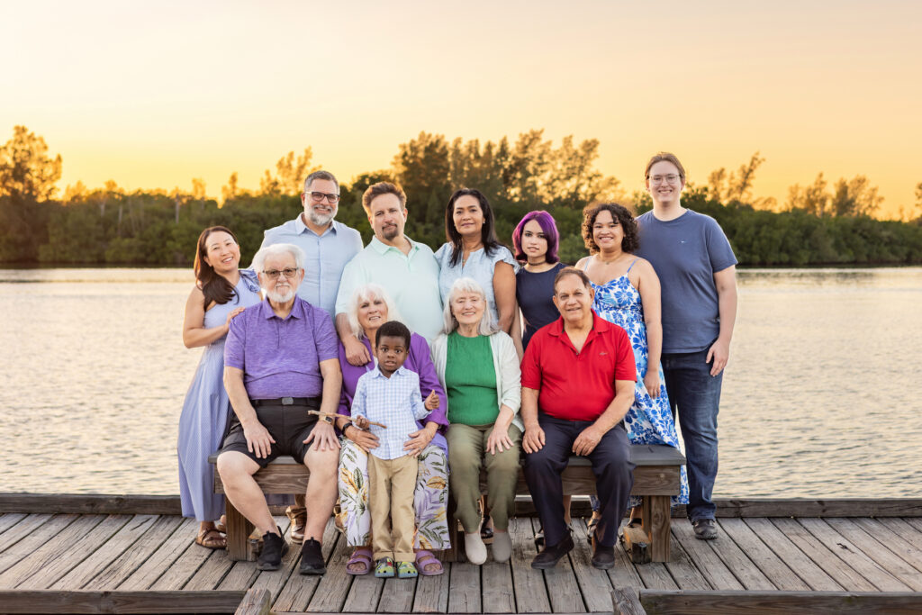 A multigenerational family poses on a wooden dock by the water at sunset, with trees in the background. Some are seated, others stand behind, and everyone is smiling. Vero Beach Photoshoot