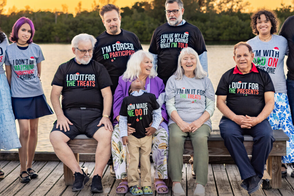 A group of people, young and old, sit and stand together on a dock by the water at sunset, all wearing humorous matching black T-shirts with funny sayings about participating in family events. Vero Beach Photoshoot