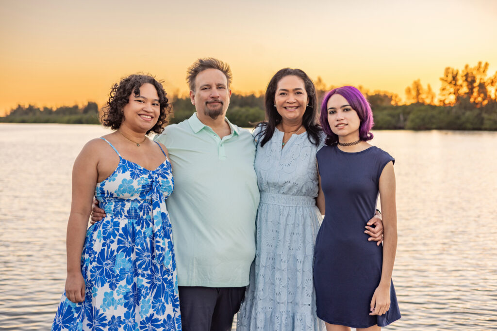 Four people stand close together, smiling by a lake at sunset. They wear summer dresses and a polo shirt. Trees are visible in the background with a warm orange sky above the water. Vero Beach Photoshoot