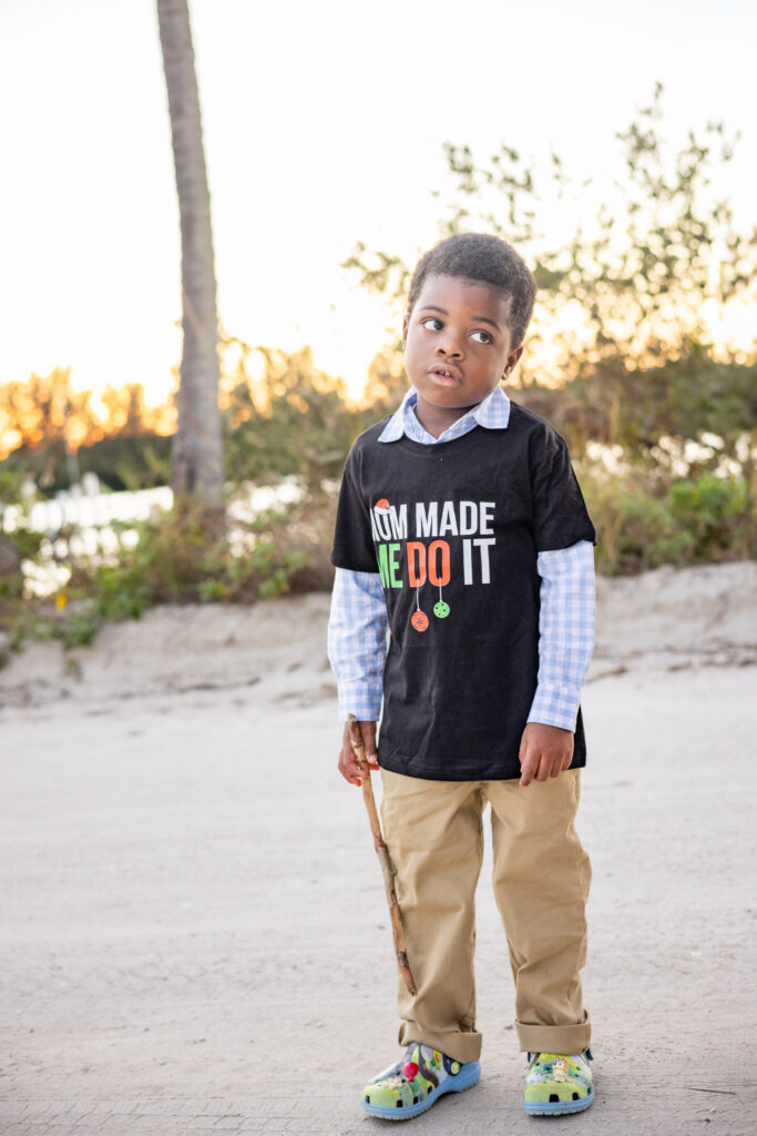 A young boy stands outdoors holding a stick, wearing khaki pants, colorful shoes, and a black T-shirt that says, “MOM MADE ME DO IT.” He looks upward with an expressive, slightly exasperated face. Vero Beach Photoshoot