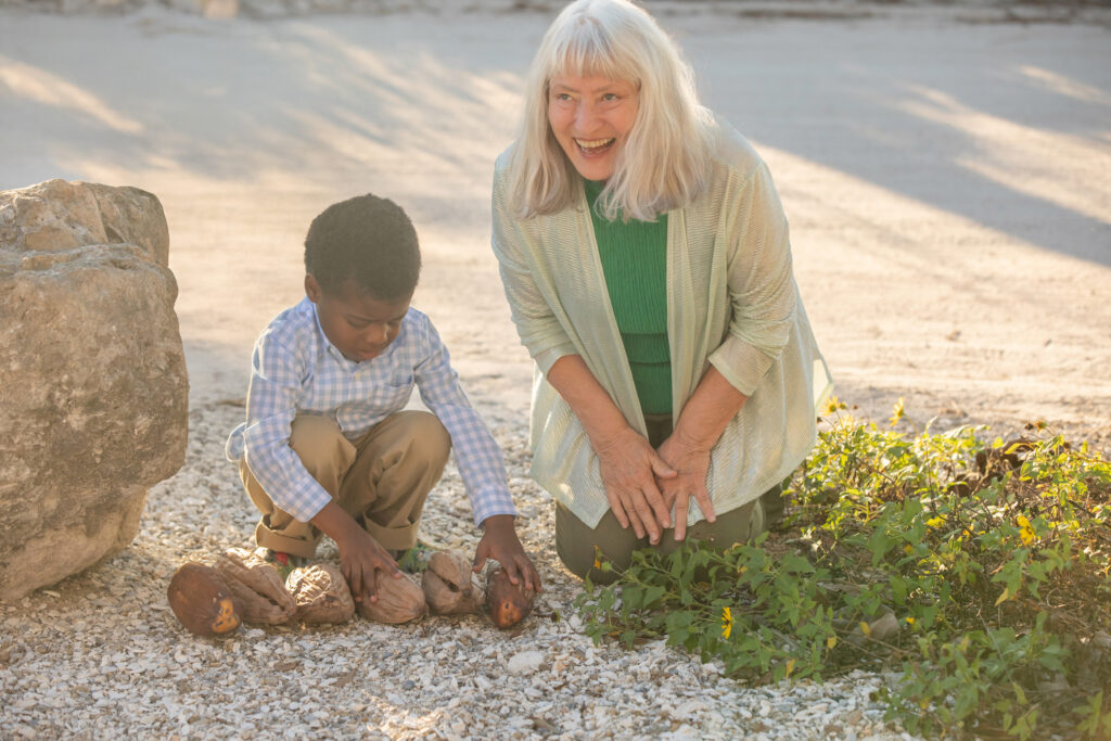 An older woman with gray hair smiles and kneels beside a young boy playing with coconuts on a gravel path near some greenery, in bright sunlight. Vero Beach Photoshoot