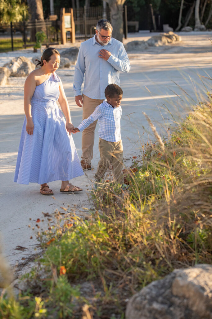 A woman, man, and young boy walk together outside on a sunny day. The woman holds the boys hand as he looks at plants by the path. They are dressed in light, summery clothes and surrounded by greenery. Vero Beach Photoshoot