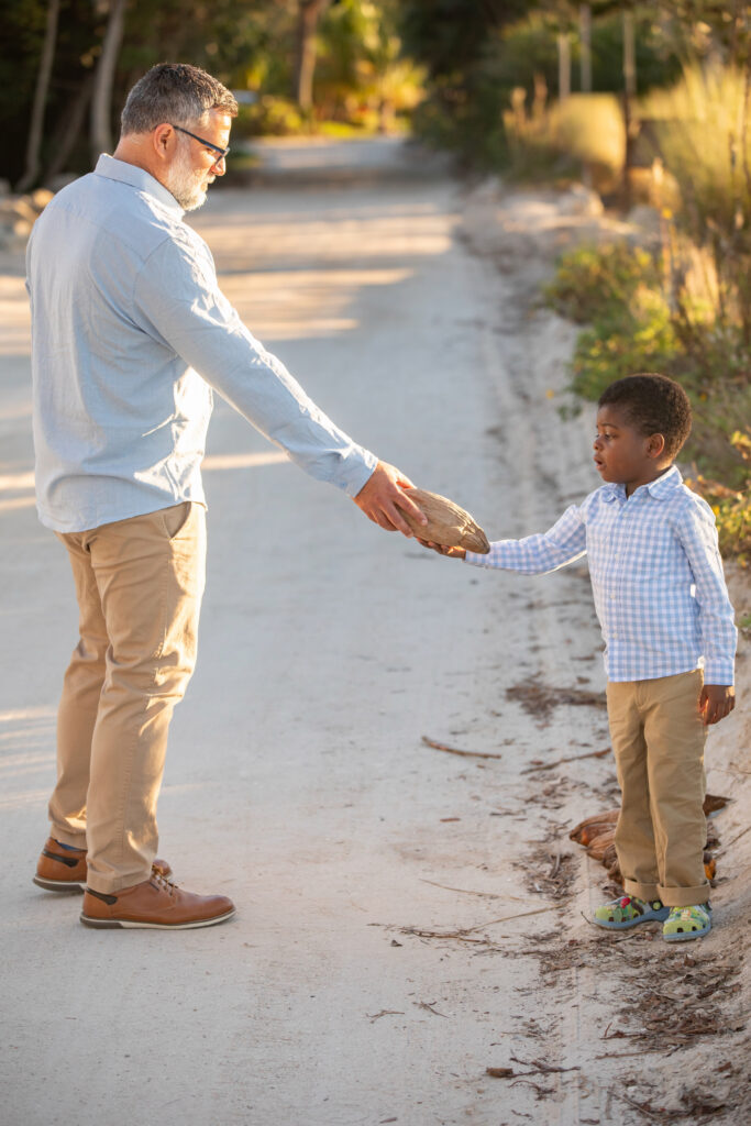 A man hands a coconut to a young boy on a sandy path surrounded by greenery. Both are dressed in light blue shirts and khaki pants, and the boy looks up at the man as he receives the coconut. Vero Beach Photoshoot