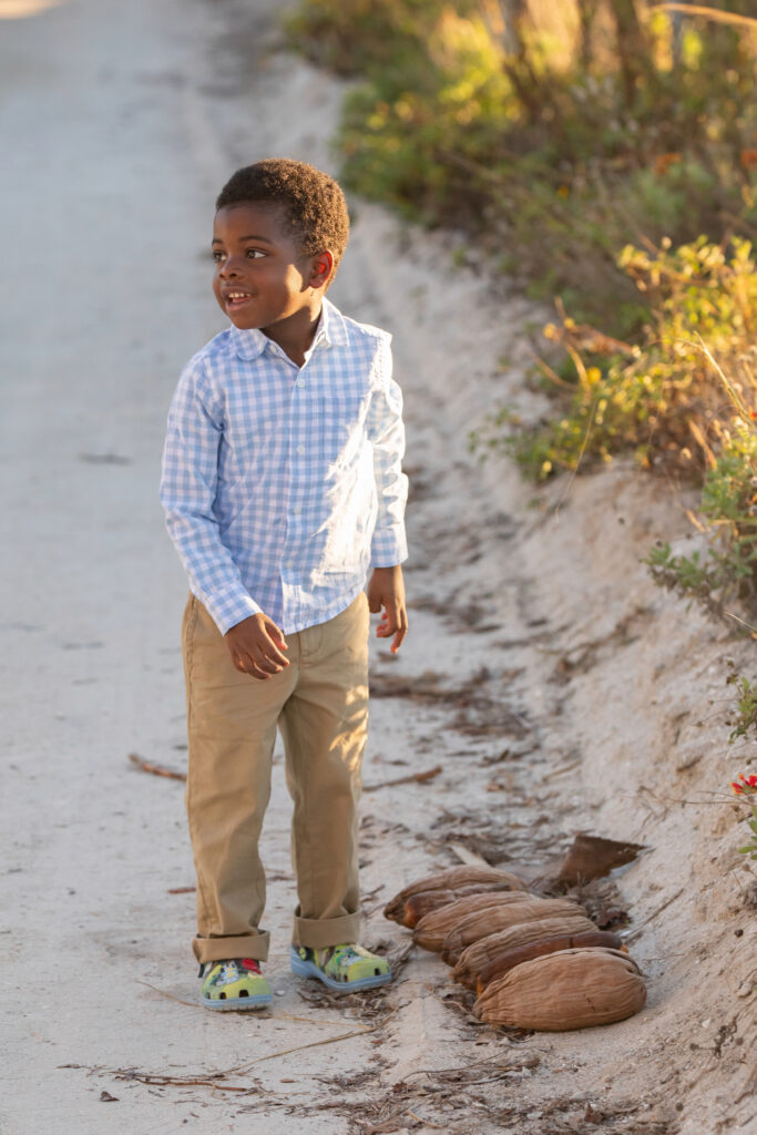 A young boy wearing a light blue checkered shirt, khaki pants, and colorful shoes stands smiling on a sunlit path next to some dried coconut husks and green plants. Vero Beach Photoshoot