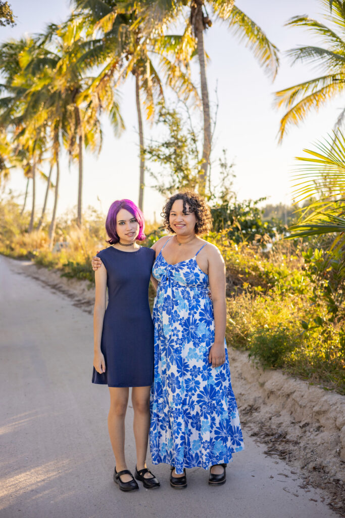 Two people stand on a sunlit path lined with palm trees. One wears a short navy dress with purple hair, the other a long blue-and-white floral dress. They smile and pose together, surrounded by greenery. Vero Beach Photoshoot