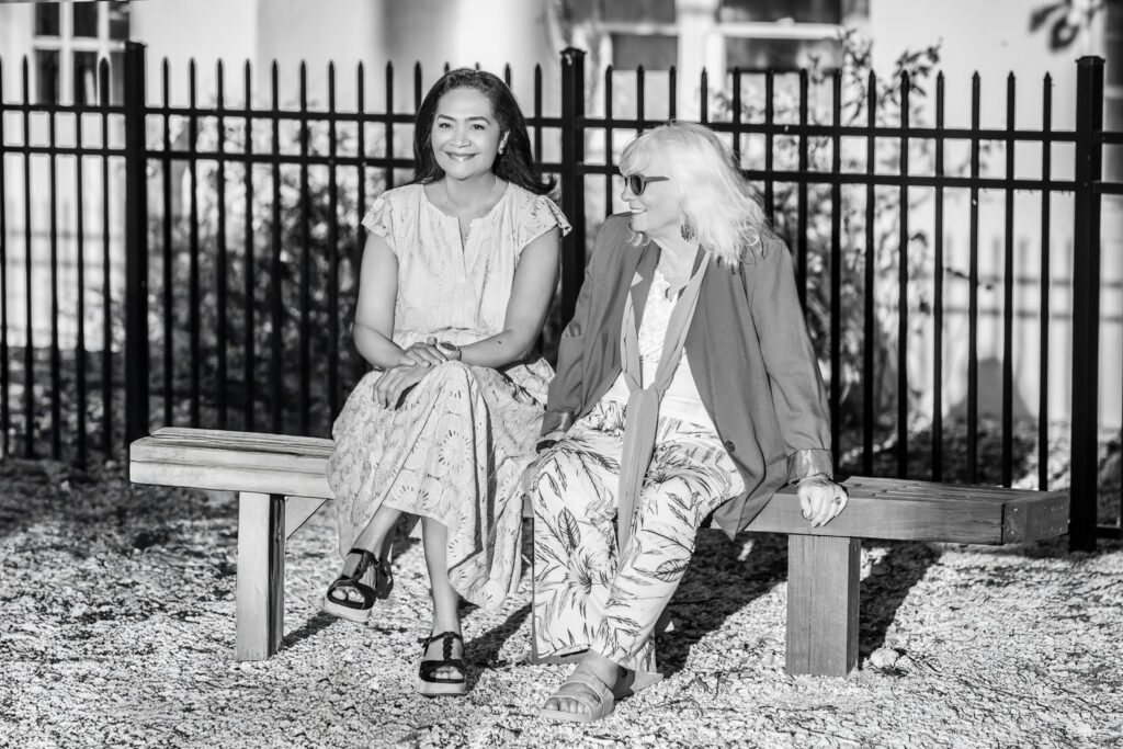 Two women sit on a wooden bench outdoors, smiling and talking. One wears a light dress and sandals; the other wears sunglasses, a light blouse, and patterned pants. A black metal fence and building are in the background. The image is in black and white. Vero Beach Photoshoot