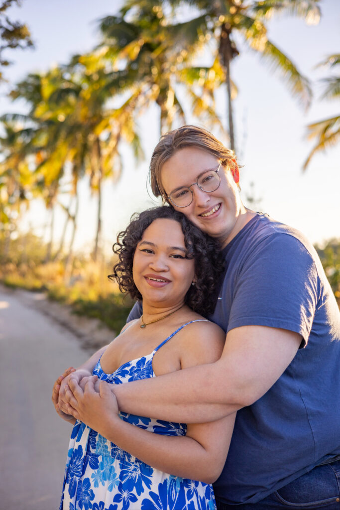 A couple stands on a sunlit path lined with palm trees. The person in front wears a blue and white dress and smiles gently, while the person behind, in glasses and a blue shirt, hugs them from behind, both looking content. Vero Beach Photoshoot