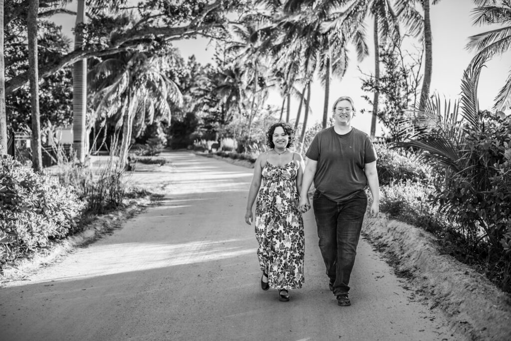 Two people walk hand in hand down a sunlit, tropical road lined with palm trees and lush greenery. One wears a floral dress, the other a t-shirt and jeans. Both are smiling and appear happy. The image is in black and white. Vero Beach Photoshoot