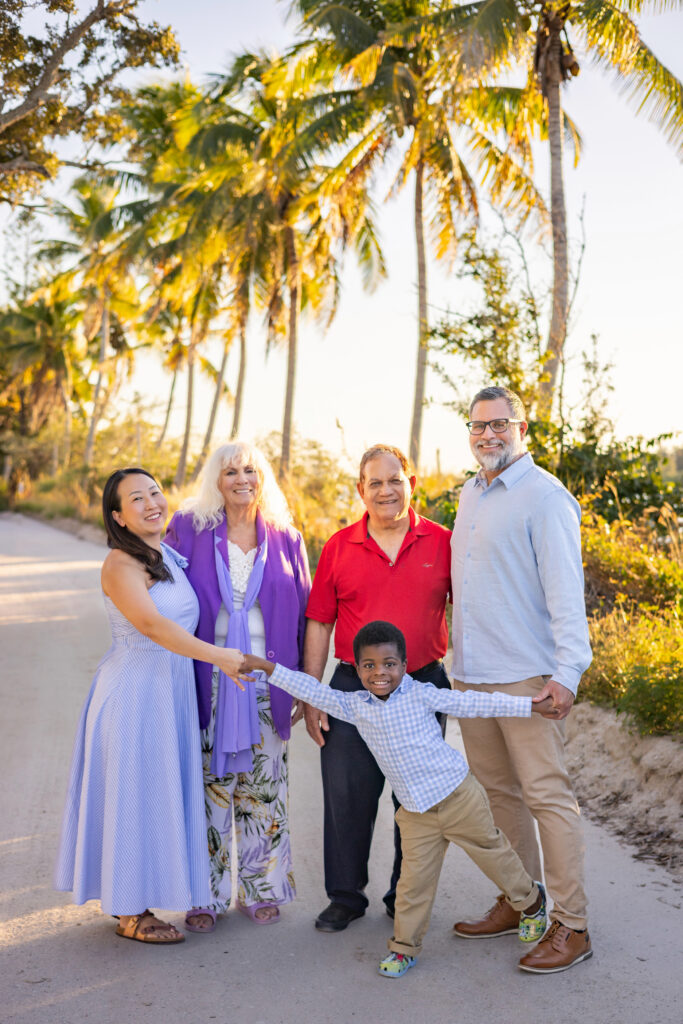 A family of five poses happily on a palm-lined path in sunlight. Two women, two men, and a young boy stand together, with the boy playfully holding hands and leaning forward. All are smiling and dressed in bright colors. Vero Beach Photoshoot