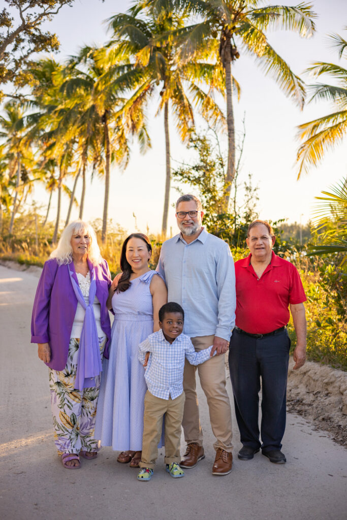 Five people stand together on a sandy path lined with palm trees, smiling at the camera in warm sunlight. Three adults, one elderly woman, one child, and a man wearing a red shirt are pictured. Vero Beach Photoshoot