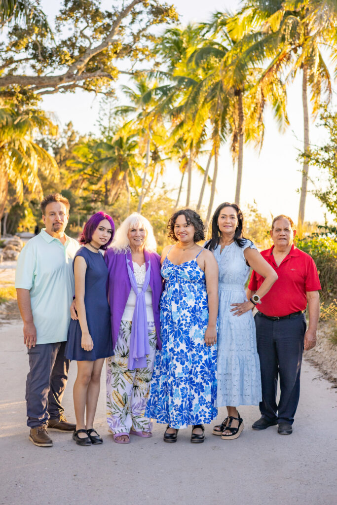 A group of six people stands together on a sunny path lined with palm trees, smiling at the camera. They are dressed in colorful, summery outfits, with greenery and the soft glow of sunset in the background. Vero Beach Photoshoot
