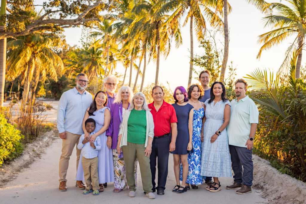 A multigenerational family of fourteen poses together on a tropical path lined with palm trees at sunset. They are dressed in casual, colorful clothing and are smiling warmly at the camera. Vero Beach Photoshoot