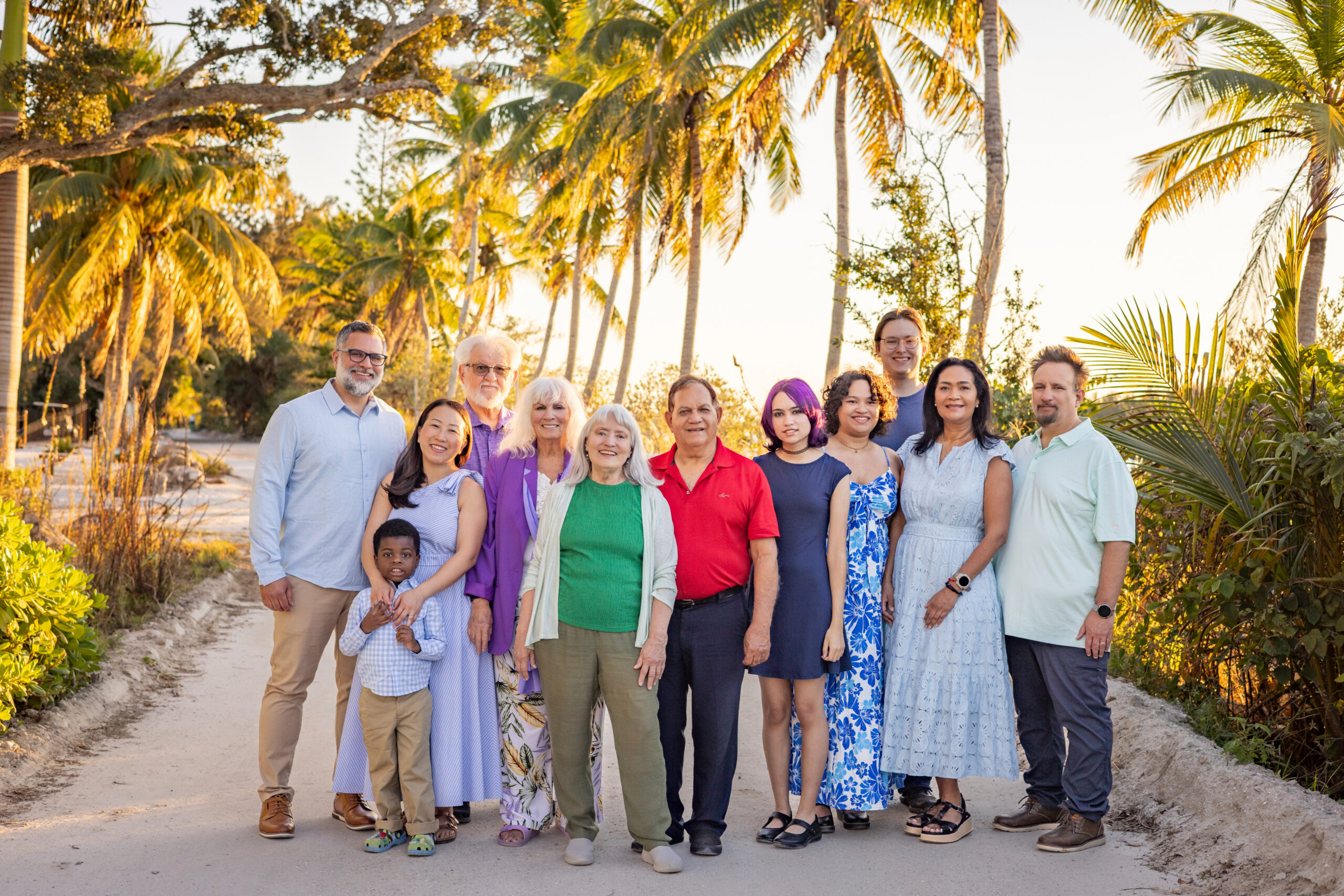 A multigenerational family of fourteen poses together on a tropical path lined with palm trees at sunset. They are dressed in casual, colorful clothing and are smiling warmly at the camera. Vero Beach Photoshoot