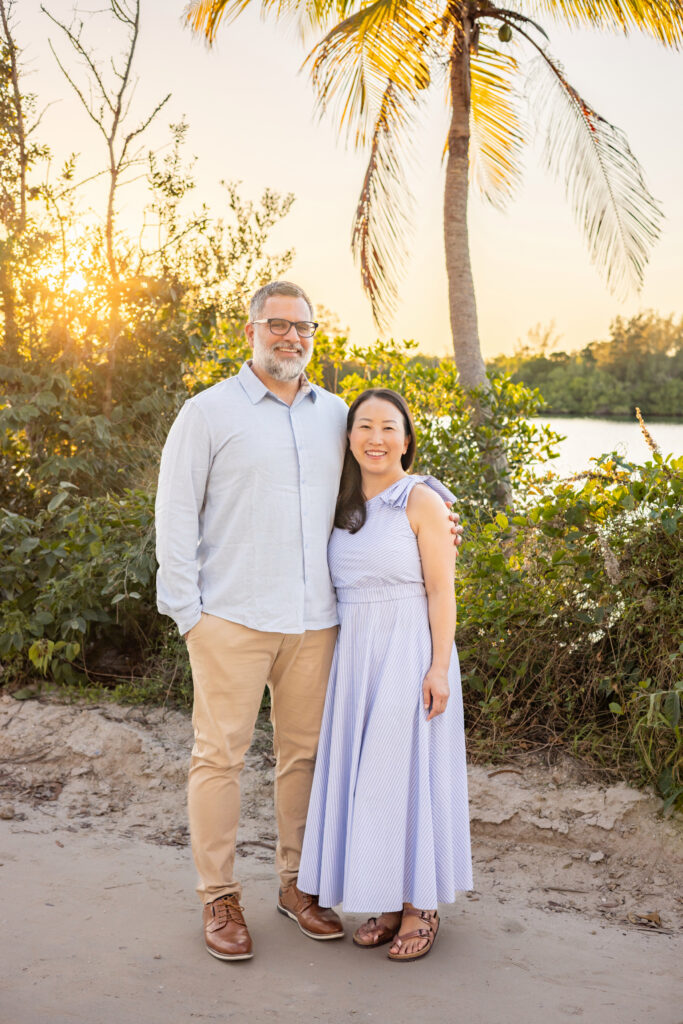 A man and woman stand close together on a sandy path by greenery and water, smiling at the camera. The sun is setting behind them, and a palm tree is visible in the background. Both are dressed in light-colored clothes. Vero Beach Photoshoot
