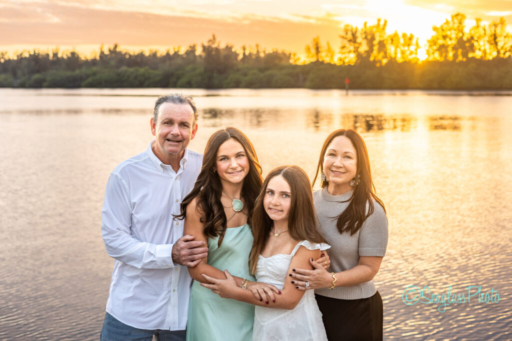 A family of four stands together by a calm lake at sunset, smiling at the camera. The parents are on each side, with two daughters in the middle. Trees and the golden sky are reflected in the water behind them. Vero Beach Photoshoot