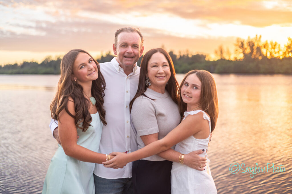 A family of four, two adults and two teenage girls, stand close together smiling by a calm lake at sunset with trees in the background, dressed in light-colored, casual clothing. Vero Beach Photoshoot
