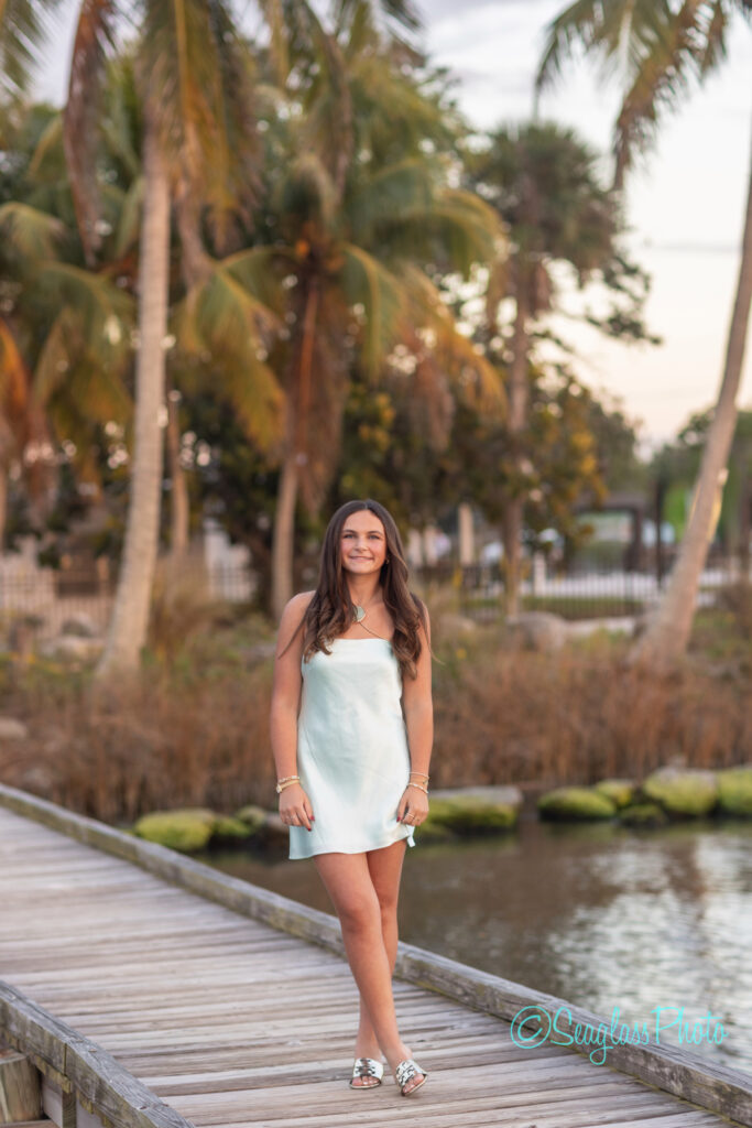 A young woman in a light blue dress stands smiling on a wooden boardwalk, with palm trees and greenery in the background on a sunny day. Vero Beach Photoshoot