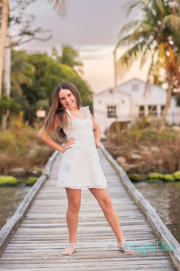 A young woman in a white dress stands smiling on a wooden dock over water, with trees and a white house in the background during sunset. Vero Beach Photoshoot